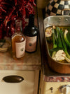 Bottles of Flamingo Estate vinegar and olive oil next to a roasting pan with garlic and leeks, set on a vintage kitchen countertop with dried red peppers in the background.