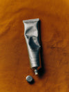 A silver tube of Flamingo Estate Manuka Rich Cream with the cap off, placed on a warm rust-colored fabric background.