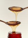 A spoon drips honey onto another lower spoon, positioned above a glass jar.