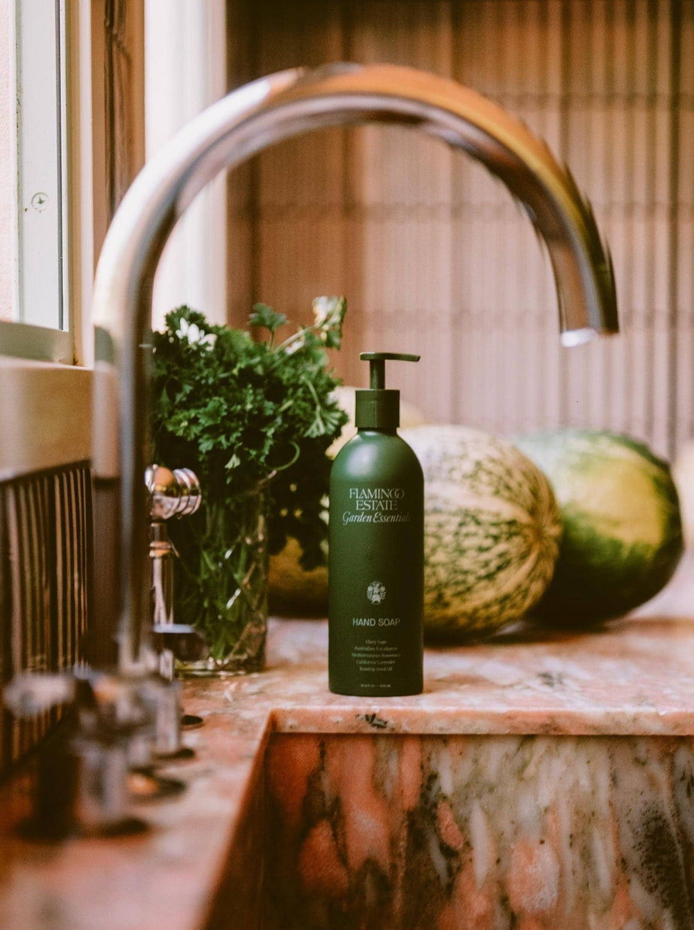 A Bottle Of Flamingo Estate Garden Essentials Handwash Behind A Sink, Next To Herbs And Watermelons