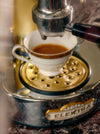 A white cup collects espresso from a metallic coffee machine.